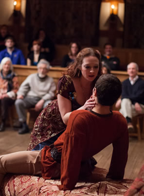 Hippolyta in flowery print dress has her hands on Arnoldo's cheeks as she leans over him on a daybed, he leaning back. Members of the audience are visible in the background.