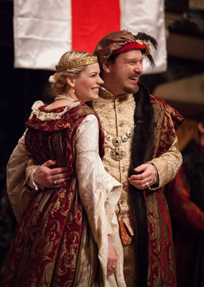 Henry with arm around Anne, both in royal clothes, standing in front a St. George Cross flag