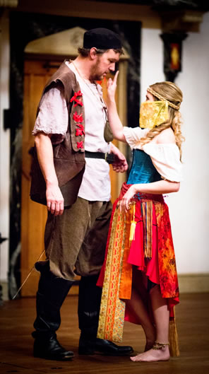 Wilmore in black hat, peasant outfit with white tunic, brown vest (featurig red ribbons)  brown pants and knee-high boots, and Hellena dressed in bright blue, red, and orange gipsy dress, yellow veil around her face, and a finger pointed at Willmore's nose.