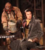 Dromio in old leather flight cap, tan work shirt and pants with vest holds up a spoon dripping white gunk into a bowl in his left hand, a man in industrial clothes, leather helmet and red goggles pours slime into another bowl from an old metal watering can, the bowl is on a table made of a metal cog on wood stand, and Antipholus sits wearing long gray overcoat, gold vest and cravat. The chairs have ladders for backs, and in the background are gears, tubes and valves on the wall