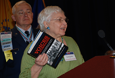 Carol Kelly in olive green dress holds "Voices of our Comrades" in her hands as she speaks at the podium as retired Air Force colonel Paul Groskreutz in his service uniform watches behind her