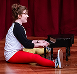 Allie Babich wearing a white t-shirt with long black sleeves and tights with one leg red and the other yellow sits on the stage playing a toy piano in The Complete Works of William Shakespeare (Abridged) [Revised] (Again).
