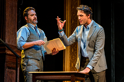 Camillo in gray three-piece suit sans jacket and holding papers in his left hand listens to Polixenes in gray jacket and unbuttoned shirt waving his right hand in consternation. 