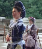 Algernon in three-piece gray suit and red striped dressing gown with hand on his and other hand across his stomach, Gwendolen in blue and white striped Victorian hat with lace trim and puffy hat, hands folded at her belly, Lady Bracknell in dress and straw hat with red bow sitting on a chair in the backround, where you see parquade floor, stone railing, and trees.