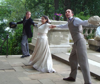 Cecily in the middle in white blouse and cream dress with arms outstretched, hands holding that of, on the far end,  Jack in black mourning coat and gray pants and holding a black tophat in his right hand, and on this end Algernon with gray vest and pants, blue shirt, and dogcap in his left hand