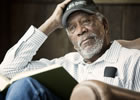 Freeman with white, close-cropped beard, wearing blue-striped white shirt, blue jeans, and a baseball cap that says "One Six Right: The Romancer of Flying", book opened on lap, right elbow resting on chair arm with hand on head, he's looking off in the distance.