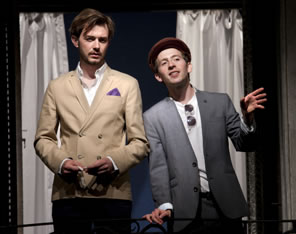 On balcony with white curtains in the background, Valentine in tan double-breasted tight-waisted dinner jacket, pocket handkerchief, dark pants, hands finger lady's white glove, Speed in open gray sport jacket, white shirt, sunglasses hanging from collar, red dog cap,  talking with right  hand on balcony rail and left hand expressing to the side.