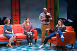 Daughter as mom  slouches in an orange chair, leg dangled over one arm, a counselor with a clipboard stands next to her, and on an orange couch sits mom as daughter straightlaced and, in similar pose, the English teacher. Red lockers in the backgroung.  