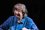 Portrait of Sir trevor Nunn, smiling, wearinga blue denim shirt against a black background